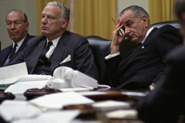 Photograph of President Johnson at an NSC meeting in 1966. He sits in the middle with two men either side. He looks bored.