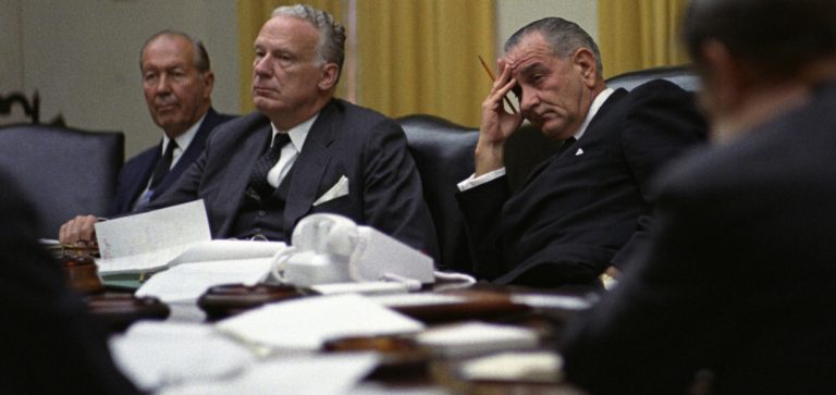 Photograph of President Johnson at an NSC meeting in 1966. He sits in the middle with two men either side. He looks bored.