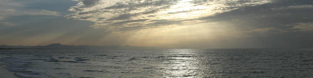 Photograph of the Breton coast, showing sky with sunlight through clouds on a grey sea with distant mountains on the horizon