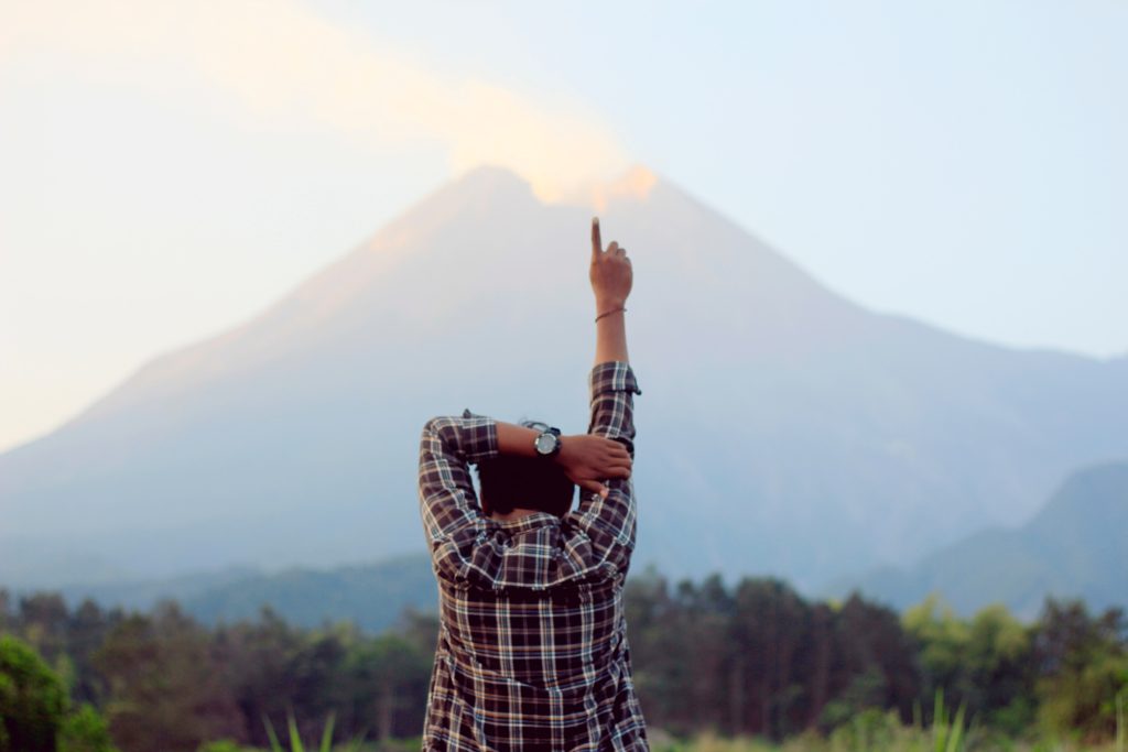 A man stands facing a smouldering volcano whilst pointing at the sky
