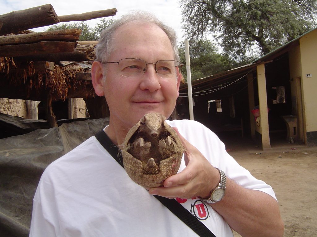 A photograph of the author Lyle Campbell during his linguistic fieldwork, Lyle is a white man with grey hair and glasses, and is holding a curled up armadillo.