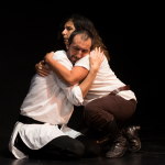 A man and woman embrace on a dark stage