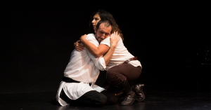 A man and woman embrace on a dark stage