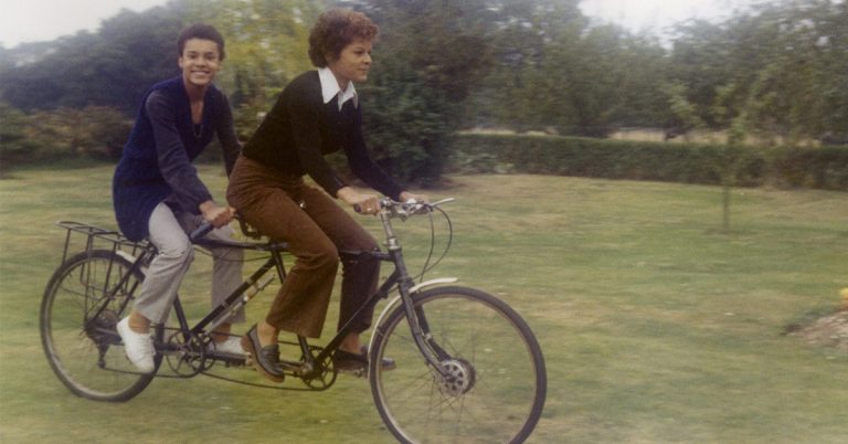 An old-time photo of two people riding on a two-seated bike across a field