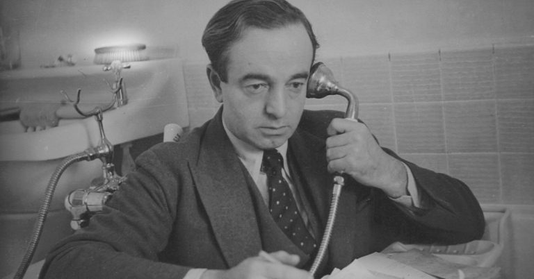 Black and white photo of a man dressed in a suit and tie sitting in a bath and holding a phone