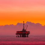 An oil rig can be seen over the horizon of the sea against a bright orange sky