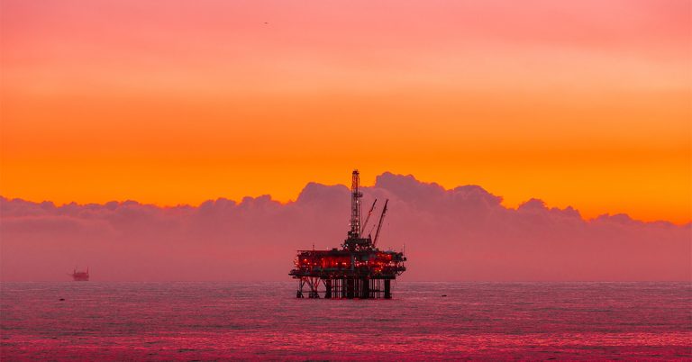 An oil rig can be seen over the horizon of the sea against a bright orange sky