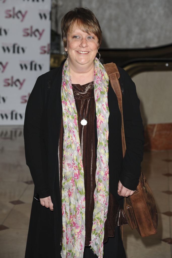 Kathy Burke smiling wearing a flowery scarf, a black coat, and brown satchel