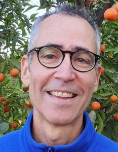 Photograph of the author, Relli Shechter. He has short greying hair, round glasses and is pictured smiling. He is set against a background of an orange tree in fruit. 