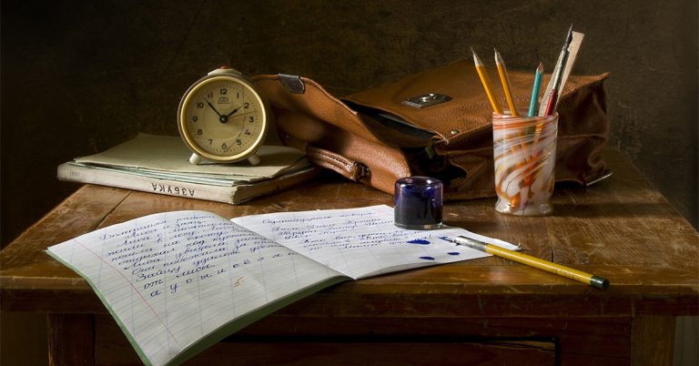 A close-up photo of a brown desk with a clock, notebook, a satchel and a cup holding a bunch of pencils