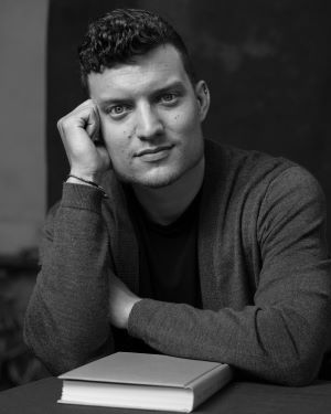 This is a black and white headshot image featuring the author Benjamin Davis looking straight at the camera, sitting at a desk with a book in front of him.