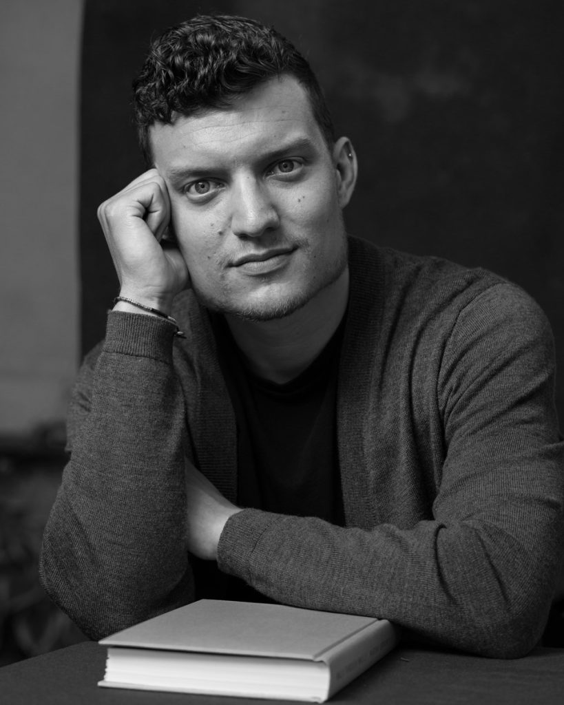 This is a black and white headshot image featuring the author Benjamin Davis looking at the camera, sitting at a desk with a book in front of him.