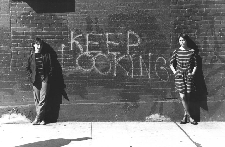 A black and white image of two women leaning against a brick wall that has the words Keep Looking written in chalk on it