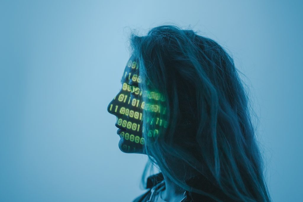 A head shot image of a female figure's profile (looking to the left hand side)  with the face being covered by projected series of digit sequences (0,1) in green colour. The background colour of the picture is light blue and the person in the image has long blond hair.