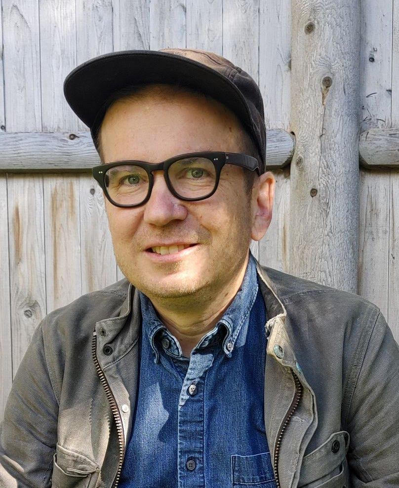 A head shot photo of Jason Read, author of 'Marx with Spinoza'. The author has green eyes, short brown hair and is bespectacled. His glasses have black frame with sharp edges. He wears a brown polo hat, a jean shirt and a grey jacket. He also smiles at the camera.