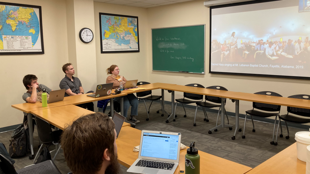 A classroom with a recording of Sacred Harp singing at Mt. Lebanon Baptist Church in Fayette, Alabama, from 2019 is broadcast on a large screen at the front of the room. Three college students are watching the recording and have their laptops out to work.