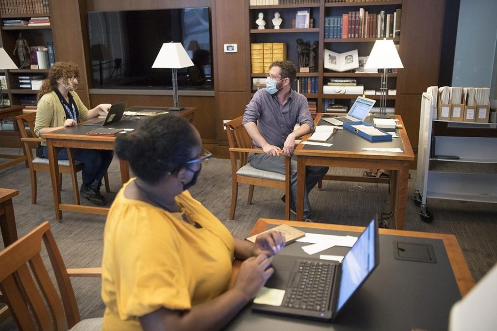 Three people sit at separate tables with laptops open, surrounded by archival papers.