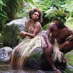A native man and woman sit on a rock in the middle of a body of water surrounded by trees