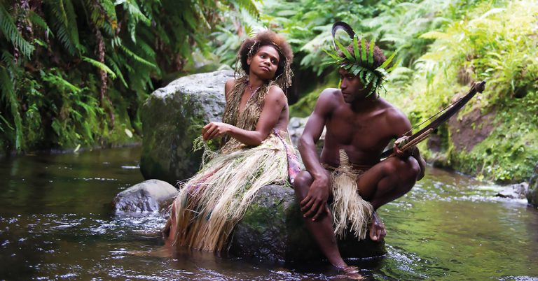 A native man and woman sit on a rock in the middle of a body of water surrounded by trees