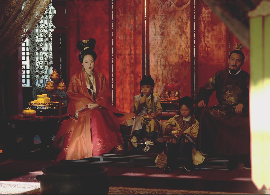 A vibrant film shot of a man and woman dressed in traditional Asian clothing beside two small children in a lavishly decorated room