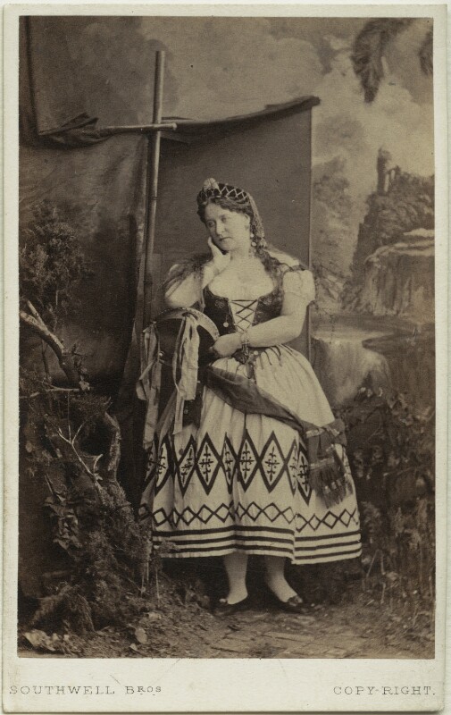 A black and white photo of Carlotta Leclercq in a corset and white dress, her hair covered in a net, and her hand raised to her cheek 