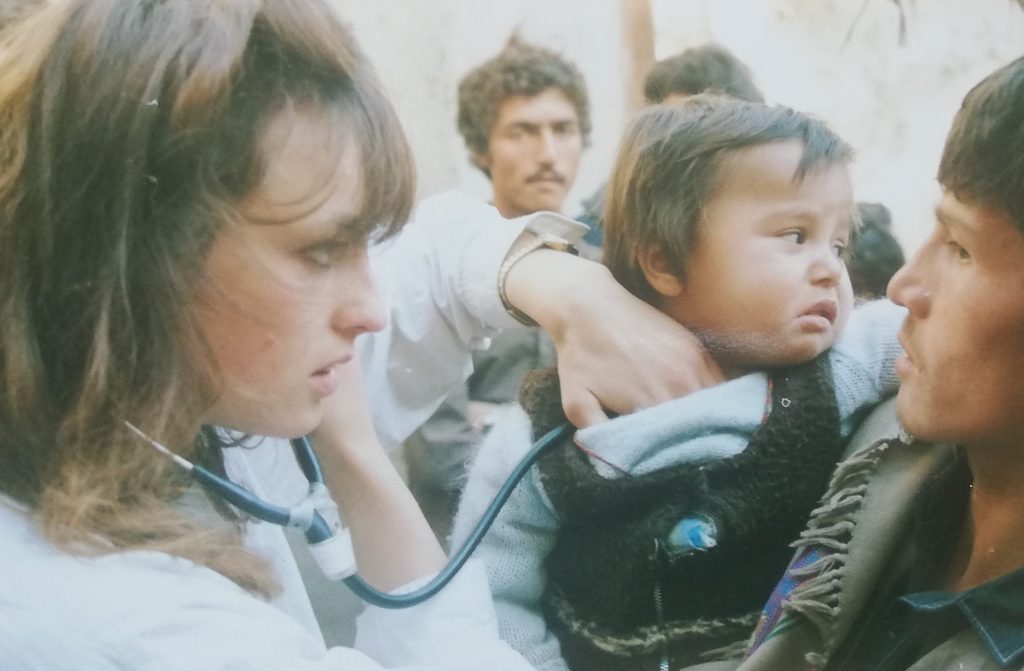 A woman with short brown hair holds a stethoscope to the chest of a small, crying child