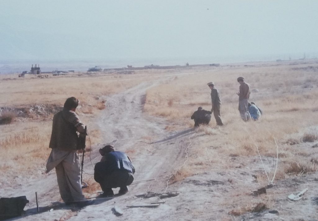 A small group of people, some standing and some kneeling down, are inspecting a dirt road for landmines