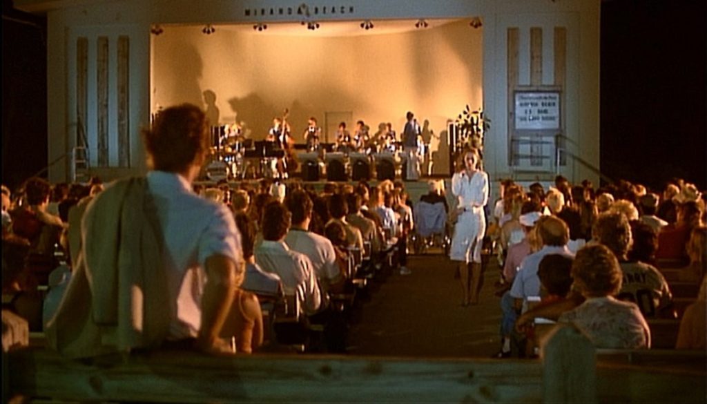 A still taken from the movie Body Heat. An audience are facing a stage watching a group of musicians. One woman walks down the centre aisle, dressed in a white dress. A man stands before her watching her. 