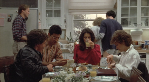 Taken from a still from the movie The Big Chill. A group of people sit around a breakfast table.  A woman in a red top sips orange juice. Three people are sitting beside her. Three more people are standing behind her around the kitchen sink.