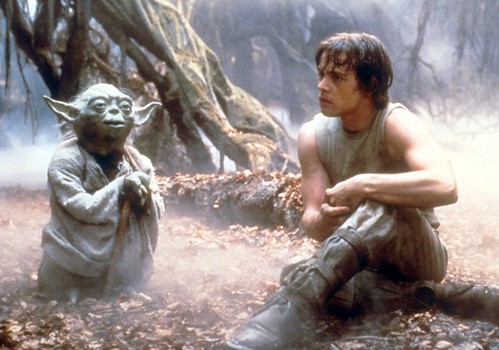 A still from the movie The Empire Strikes Back. A man sits in a clearing in the woods talking to Yoda, a short green creature.
