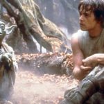 A man with curly hair sits in a forest clearing next to Yoda, a green elf like creature with pointy ears