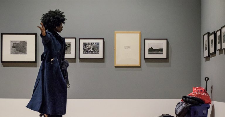 A woman stands to her side extending both arms horizontally in a room lined with picture frames. She is standing in front of a shopping cart filled with trash bags.