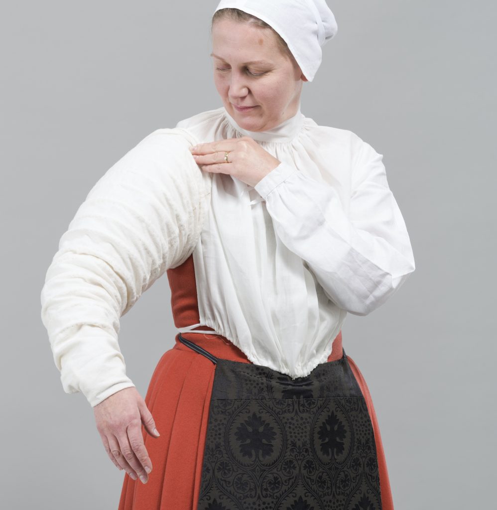 A woman dressed in period clothing and a white bonnet demonstrates putting on a farthingale sleeve