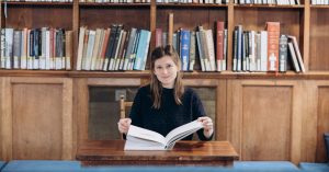 Librarian Eve Lacey sits at a desk with an open book, there are shelves of books in the background