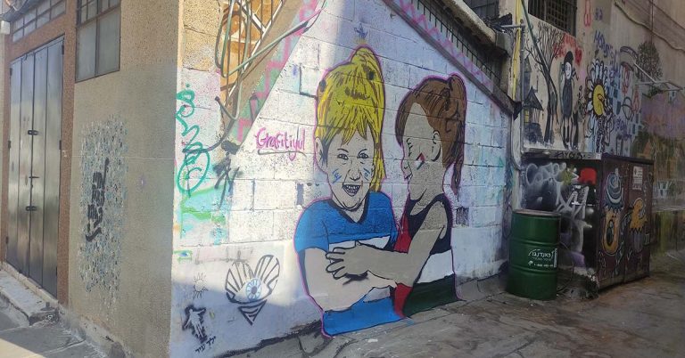 A wall is covered in a graffiti mural of two young girls laughing together, one wears the colours of the Palestinian flag, the other wears the colours of the Israeli flag