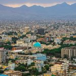 Panorama photograph showing a densely built, colourful city set against a mountain backdrop