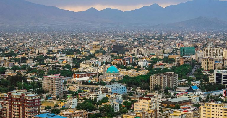 Panorama photograph showing a densely built, colourful city set against a mountain backdrop