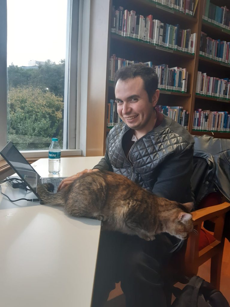 A man sits in front of a laptop and smiles at the camera, with a cat sat on the desk next to him.