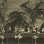 Black and white image of 13 women sitting at a conference-style table. One holds a banner with text reading 'Un pour tous, Tous pour un' - the French for 'one for all, all for one'.