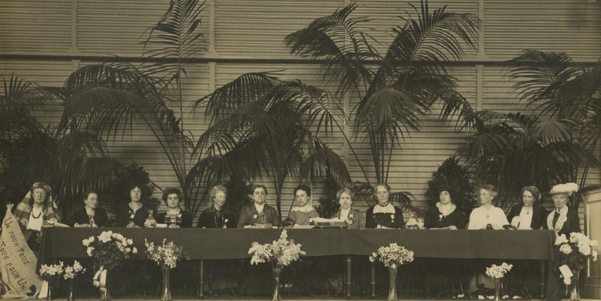 Black and white image of 13 women sitting at a conference-style table. One holds a banner with text reading 'Un pour tous, Tous pour un' - the French for 'one for all, all for one'.