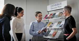 You can see four people in conversation, one holding a book, all standing in front of a book display with the banner 'Edinburgh University Press'. This is representative of EUP staff.