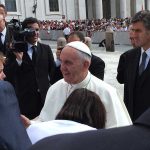 Pope Francis greets people at the Vatican, dressed in white papal robes and surrounded by men in suits