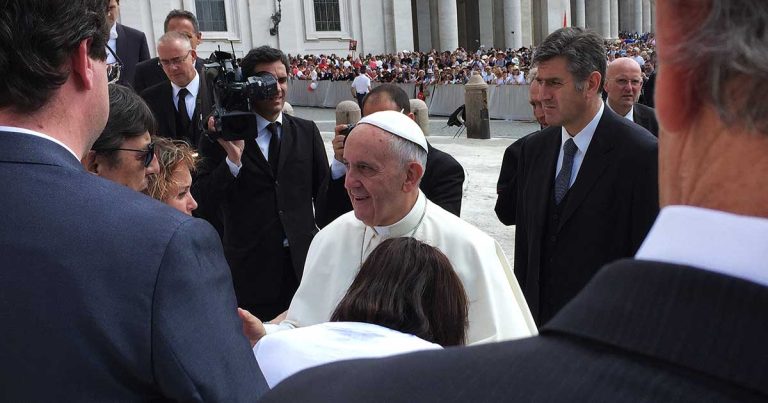 Pope Francis greets people at the Vatican, dressed in white papal robes and surrounded by men in suits