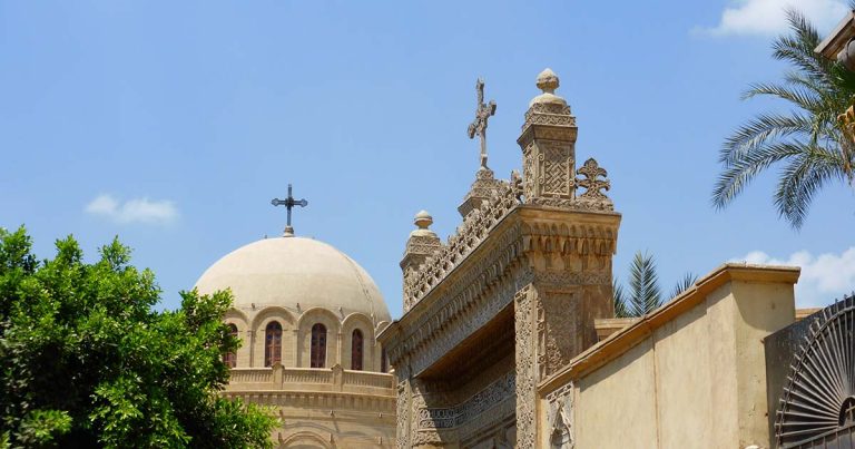 A photograph of a domed building and an elaborate archway, both with crosses on top, set against a blue sky