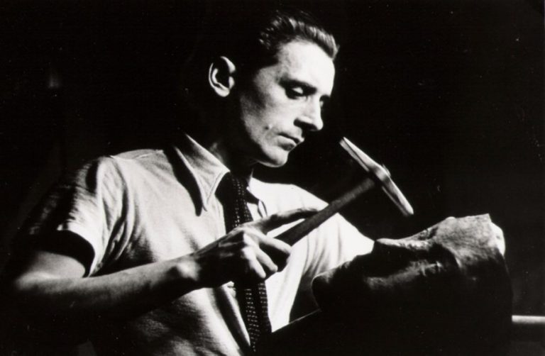 A black and white photograph of Hermann Gross holding a metalworking tool, with an in-progress sculpture before him.