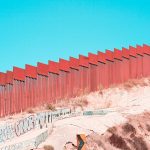 a red wall stretches along the US-Mexico border, with a sandy hill in front and blue sky behind