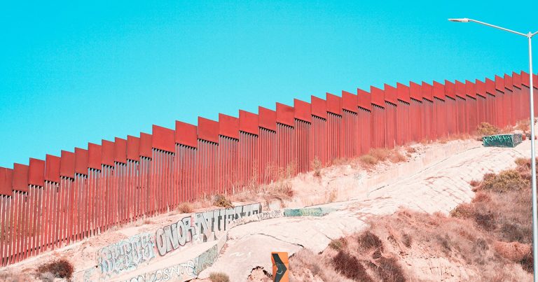 a red wall stretches along the US-Mexico border, with a sandy hill in front and blue sky behind