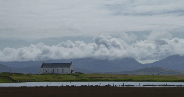 Landscape view of a remote house, with water in the foreground and mountains in the background.