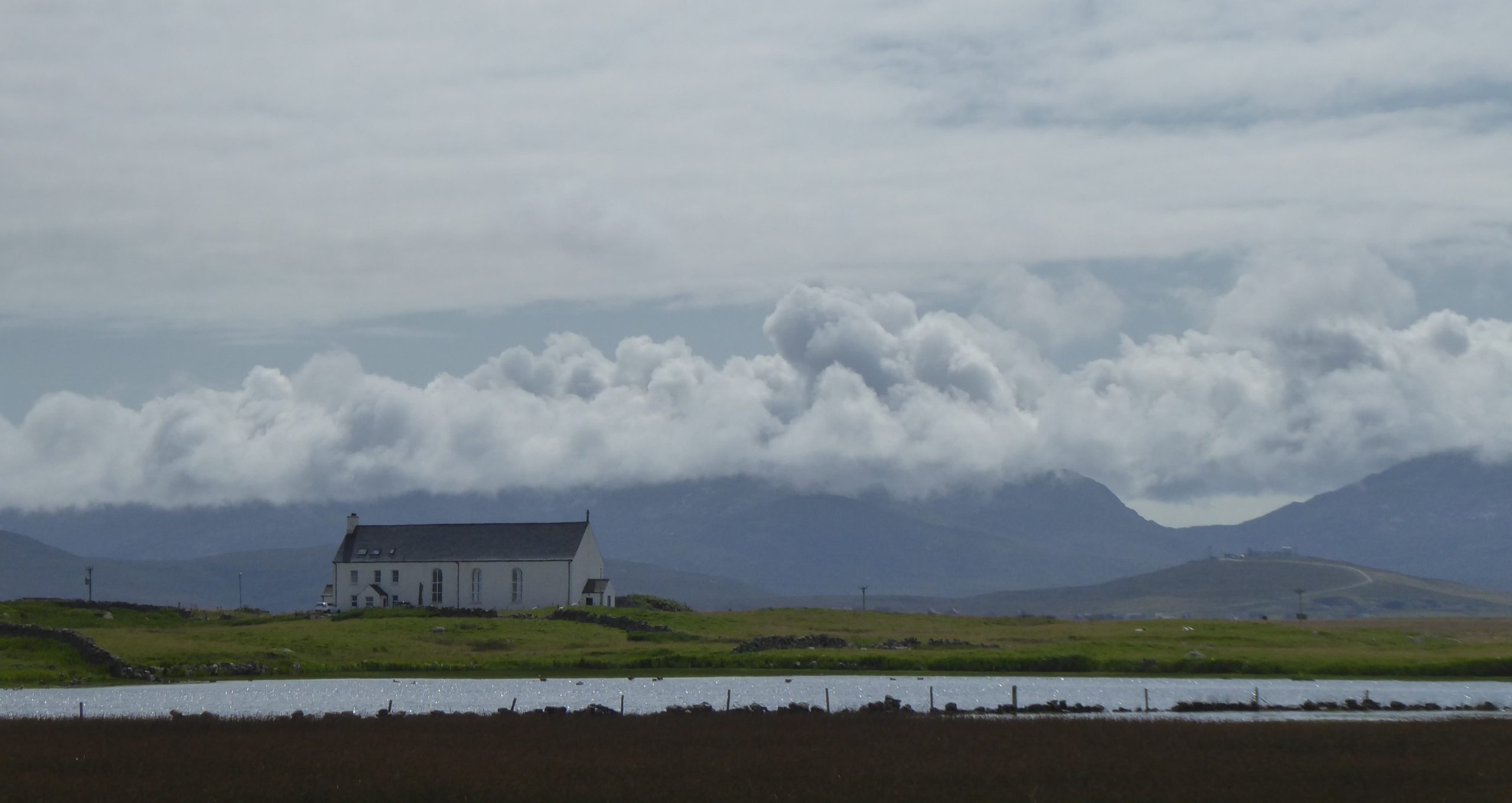 Landscape view of a remote house, with water in the foreground and mountains in the background.