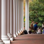 Ryan J. Johnson sits on a covered walkway, surrounded by students, with columns stretching to the left.